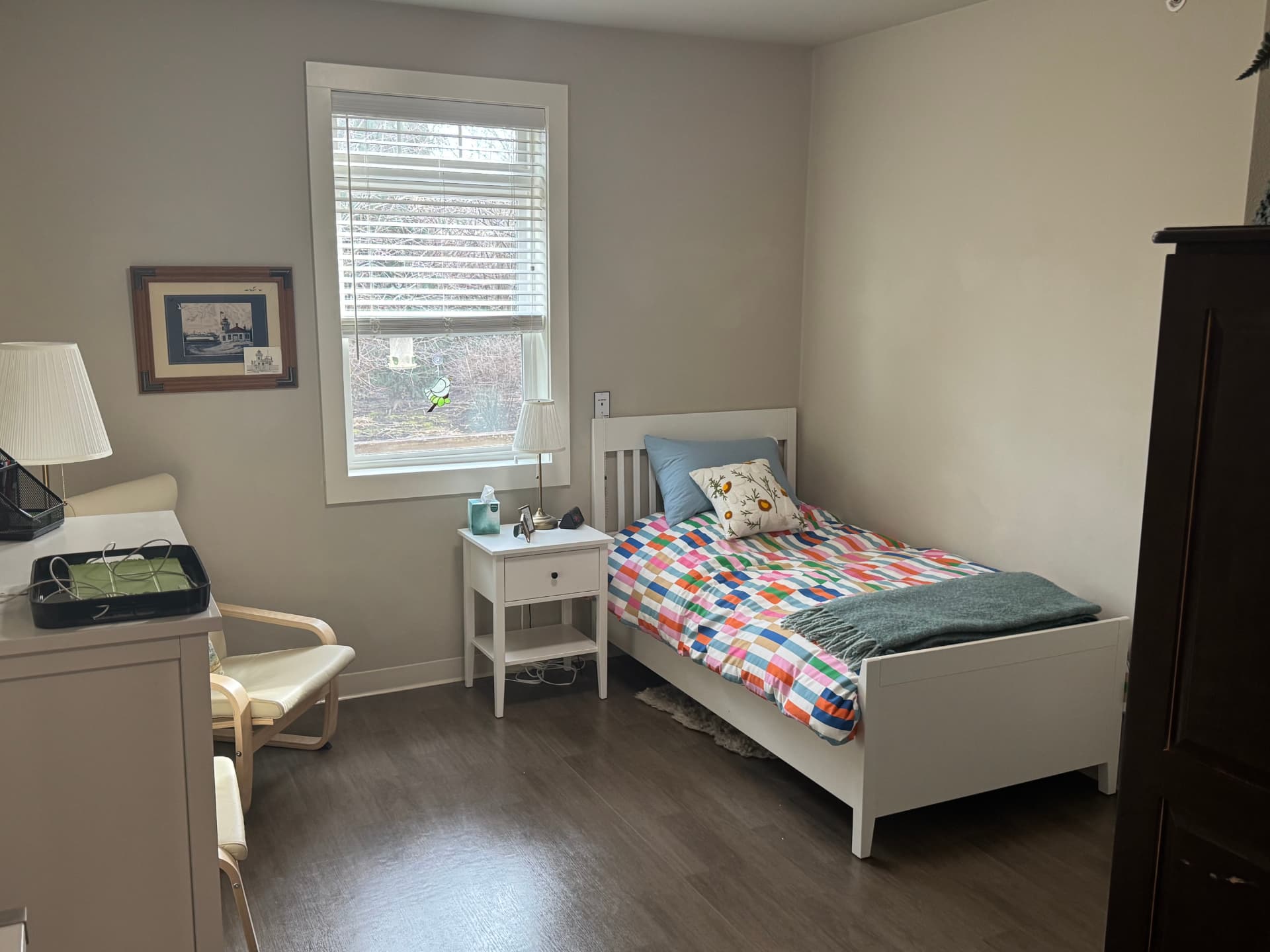 Furnished resident room with a single bed under a checked quilt and a green throw, a white nightstand with a lamp, a window with blinds, a small armchair and a dresser