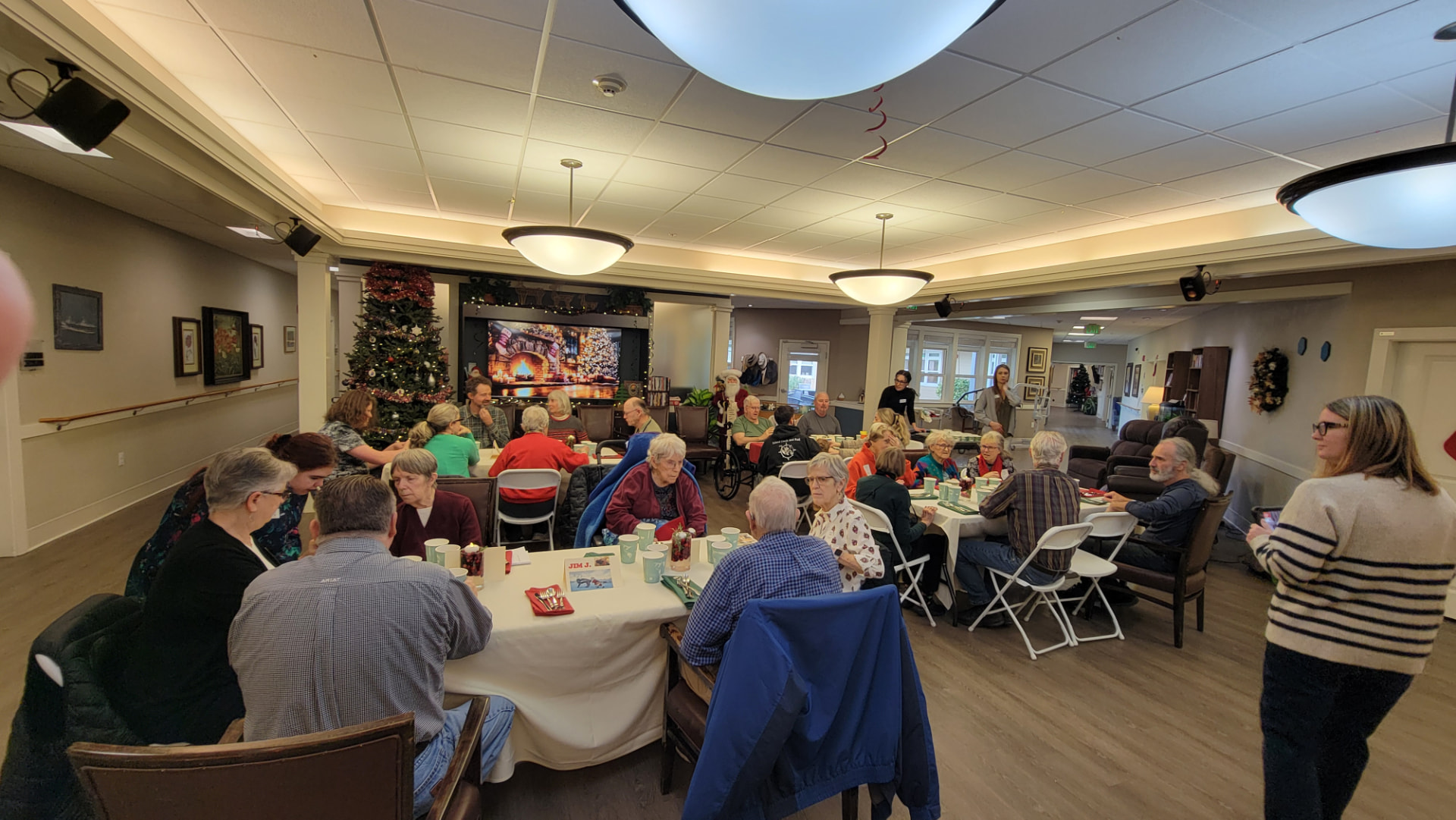 Holiday gathering in the community room, with a decorated Christmas tree at the left, a wreath on the wall, and people seated around tables covered in white cloths and set with light blue cups and plates