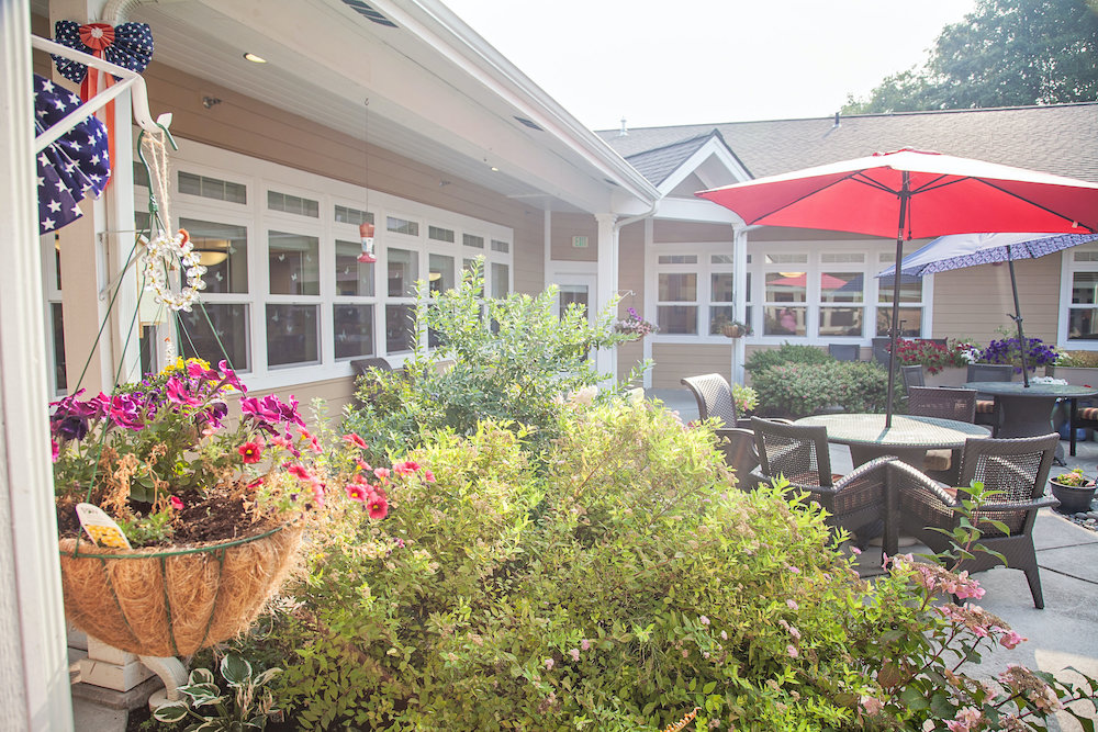 Courtyard at Lighthouse Memory Care seen past a hanging basket of pink and red flowers and flag bunting, with wicker chairs under a red umbrella beside the windows