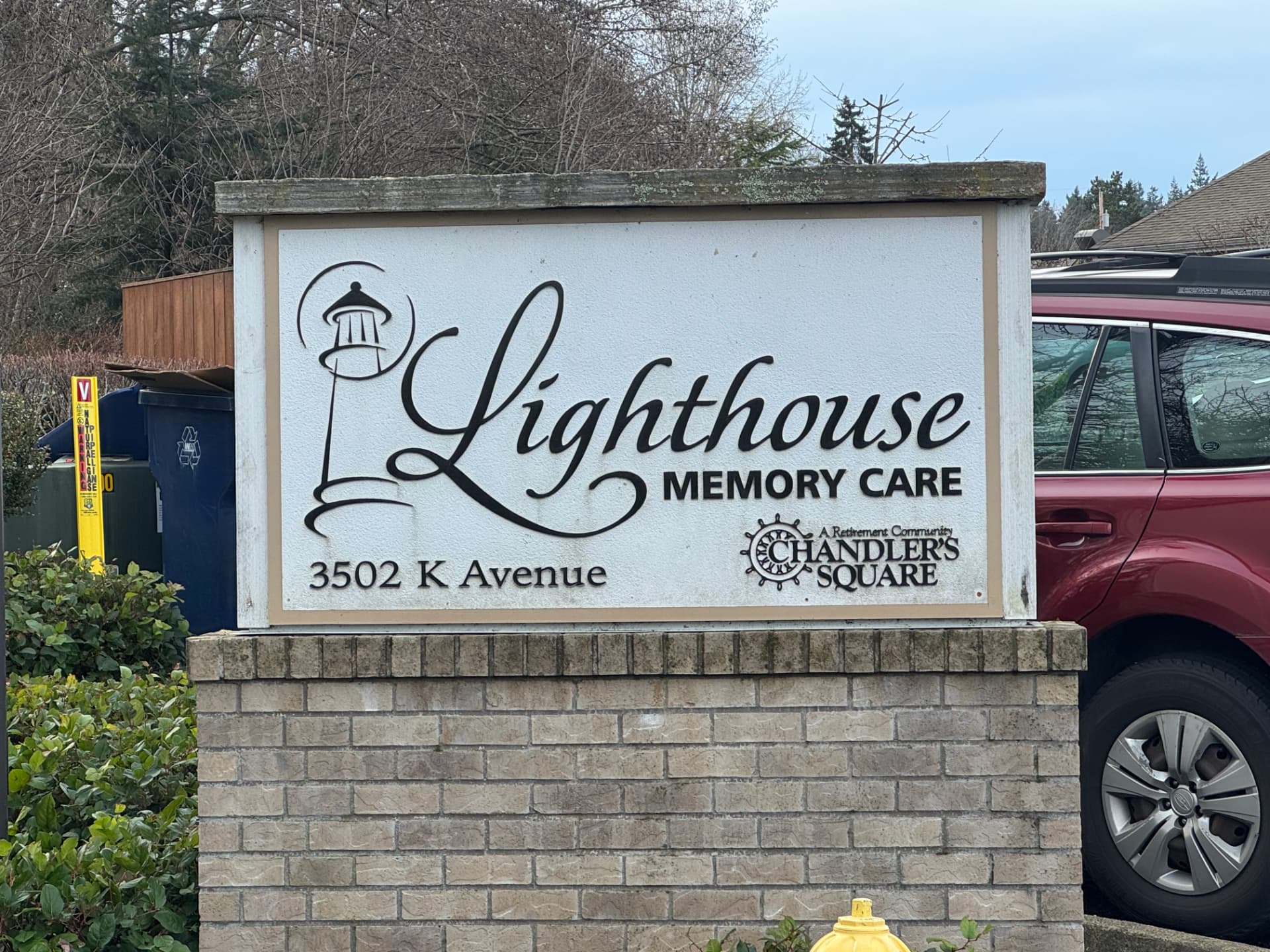 Street entrance sign for Lighthouse Memory Care, a white panel with a black lighthouse drawing and the address 3502 K Avenue, set on a brick base with shrubs in front and bare trees behind