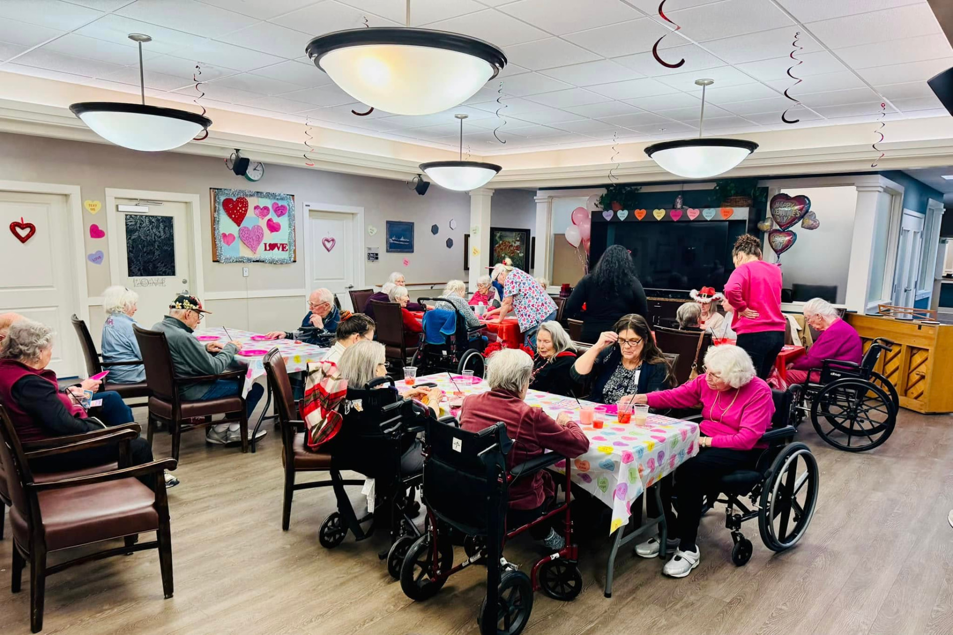 Residents seated at tables with candy heart tablecloths during a Valentine's event, paper hearts on the walls, heart balloons and hanging streamers overhead