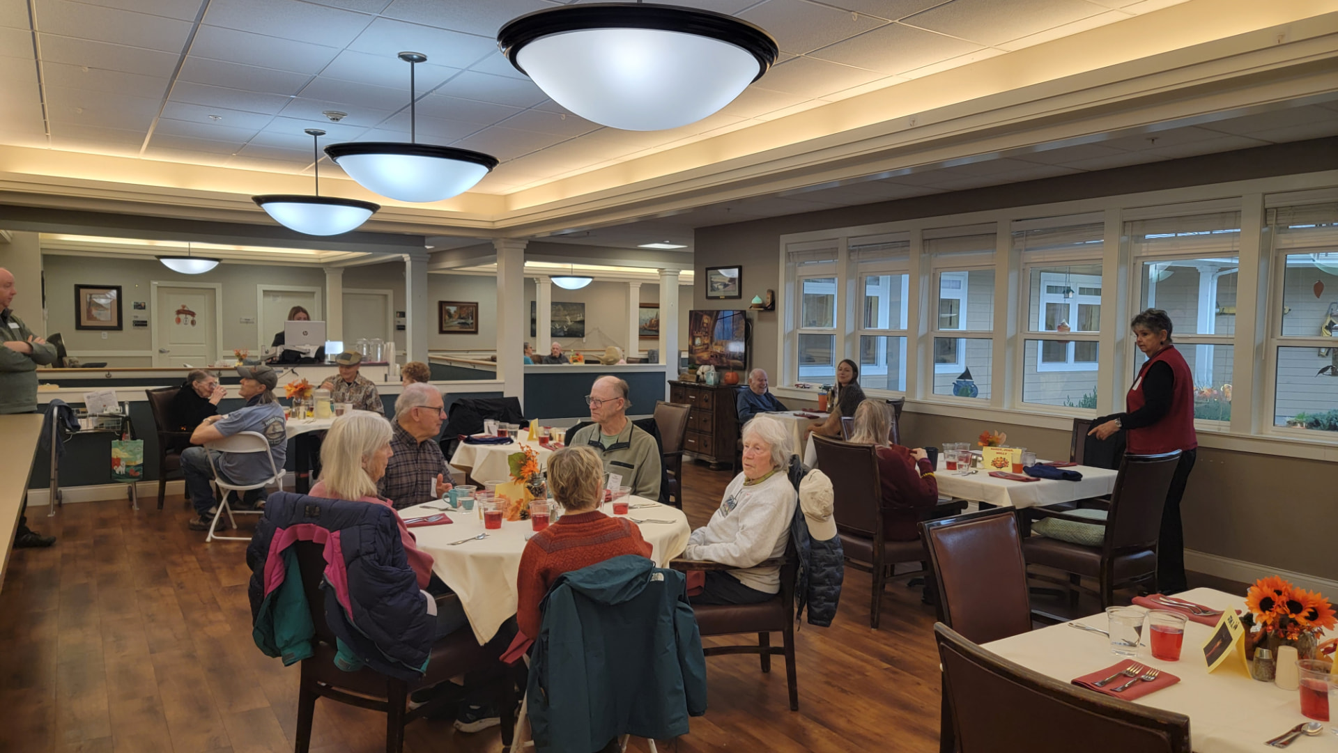 Residents seated at round tables with white cloths in the dining room, autumn leaf centrepieces and glasses of juice set out, tall windows along the far wall and three domed pendant lights overhead