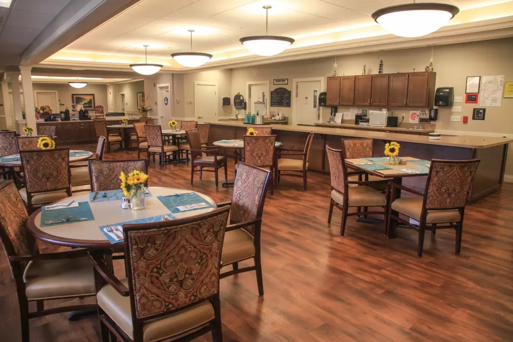 Dining room at Lighthouse Memory Care with round tables set with blue placemats and small vases of sunflowers, upholstered chairs and a serving counter along the far wall